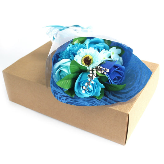 Bouquet of blue and turquoise artificial roses and blooms wrapped in blue tissue sits atop a plain kraft cardboard gift box on a white background.