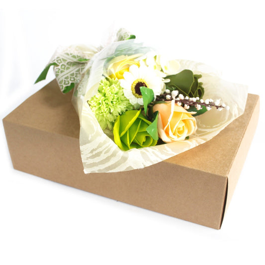 A small bouquet rests atop a closed brown cardboard box, wrapped in translucent patterned paper showing peach and green roses, a white daisy, greenery, and a lace ribbon.