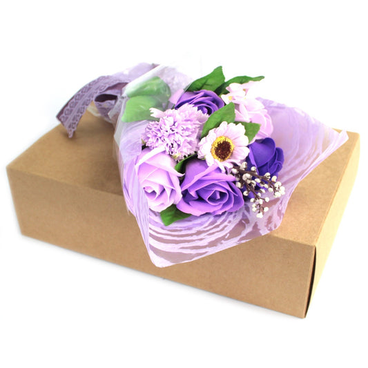 A small bouquet of lavender and purple artificial roses wrapped in purple cellophane and ribbon rests atop a plain brown cardboard box against a bright white background.