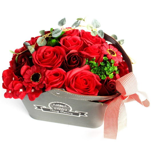 Bouquet of red roses and mixed blossoms nestled inside a gray leather-handled box labeled "Soap Flowers" with a red gingham bow sitting on a white background.