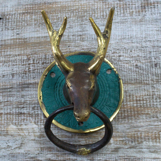 A brass deer head door knocker, affixed to a teal circular plate, is mounted on a light-colored wooden surface.