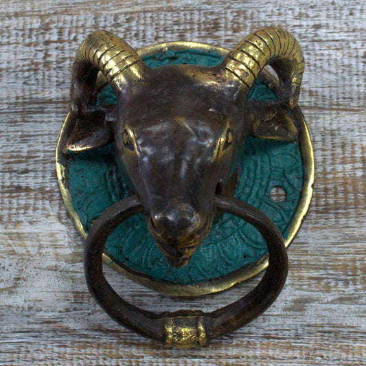 A ram-head door knocker, fashioned from dark metal with gold accents, is mounted on a teal circular plate affixed to a light-colored wooden surface.