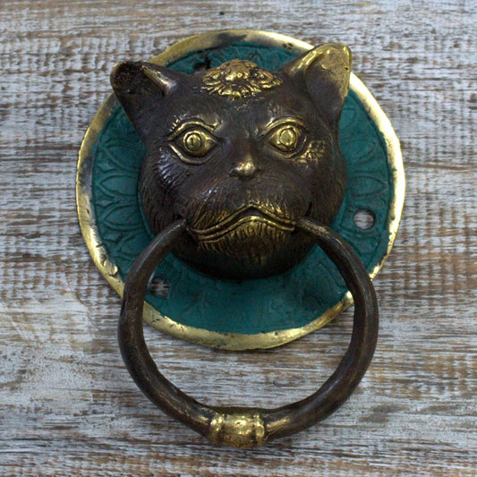 A bronze cat-shaped door knocker is mounted on a teal-green circular plate. The knocker is affixed to a wooden surface.