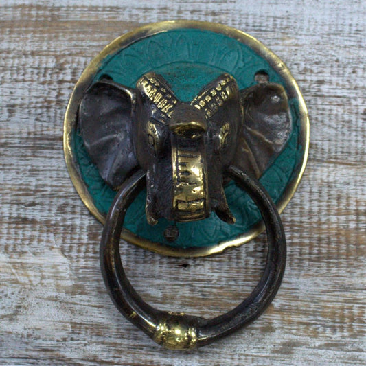 An elephant-shaped door knocker hangs on a teal and gold circular plate; it is mounted on a rustic, whitewashed wooden surface.