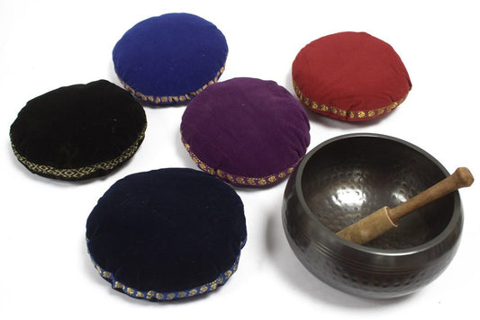 Five colored round cushions lie on a white surface, surrounding a dark metal singing bowl with a wooden mallet resting inside. The cushions have ornate, patterned trim.
