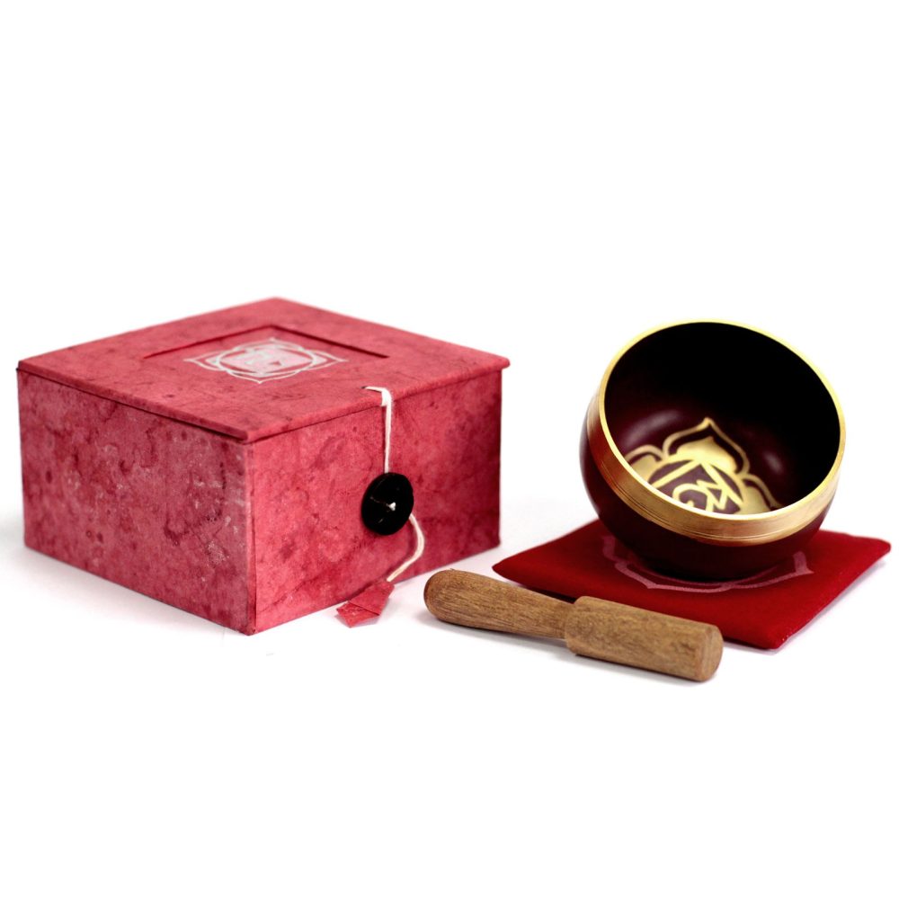 A small brass singing bowl sits tilted on a red cushion with engraved design, accompanied by a wooden mallet, next to a red decorative box with a closure button.