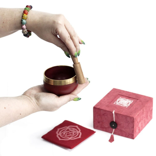 A person holds a small red and gold singing bowl with a wooden mallet. Nearby are a decorative red box and cloth with a rose design, set against a plain background.
