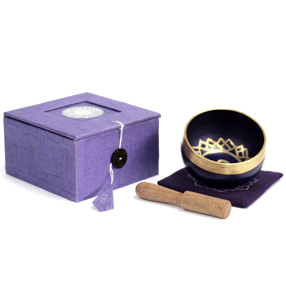 A Tibetan singing bowl with intricate gold detailing rests on a purple cushion. Accompanied by a wooden mallet, it sits beside a matching purple decorative box.
