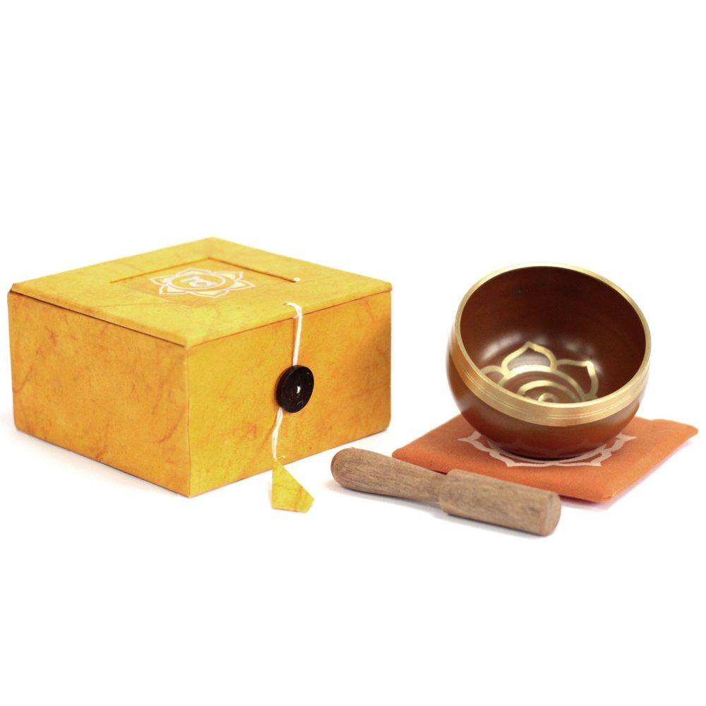A brass singing bowl rests on an orange cushion, accompanied by a wooden mallet. Nearby, a closed yellow box with white and black design elements enhances the serene setting.