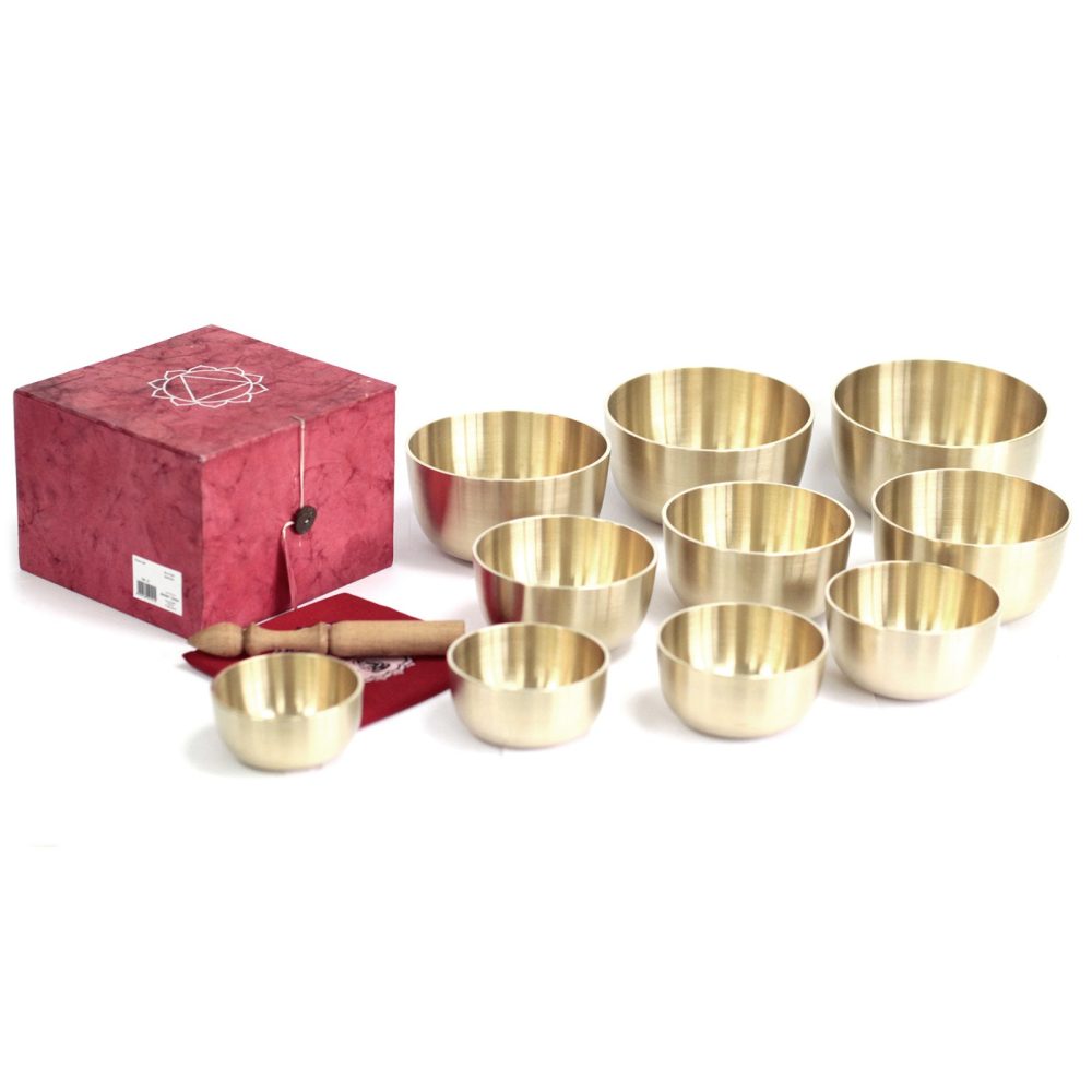 Brass singing bowls are arranged in a cluster, accompanied by a wooden mallet and red cushion. A red box with a geometric design sits nearby on a white surface.