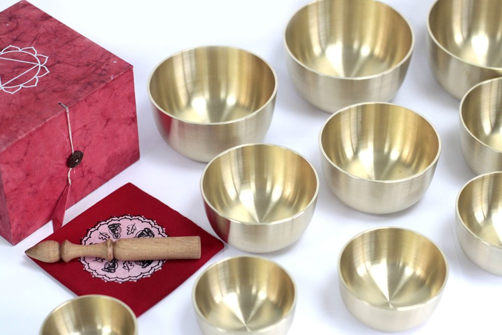 Brass singing bowls arranged beside a red box and cloth, with a wooden mallet resting on the cloth. The setting is minimalist with a white background.