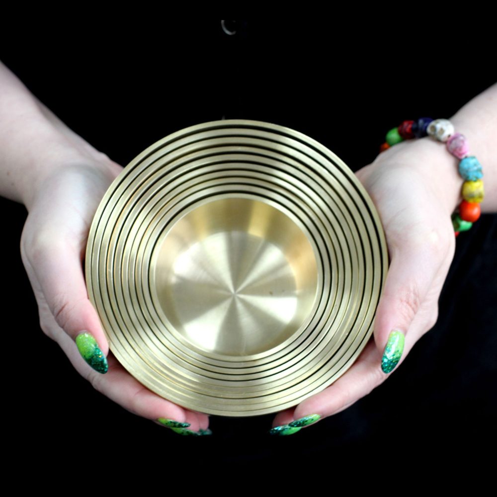 Hands hold a stack of nested, gold-toned concentric metal bowls. The person wears a colorful bracelet and has green-painted fingernails. The background is a dark, neutral surface.