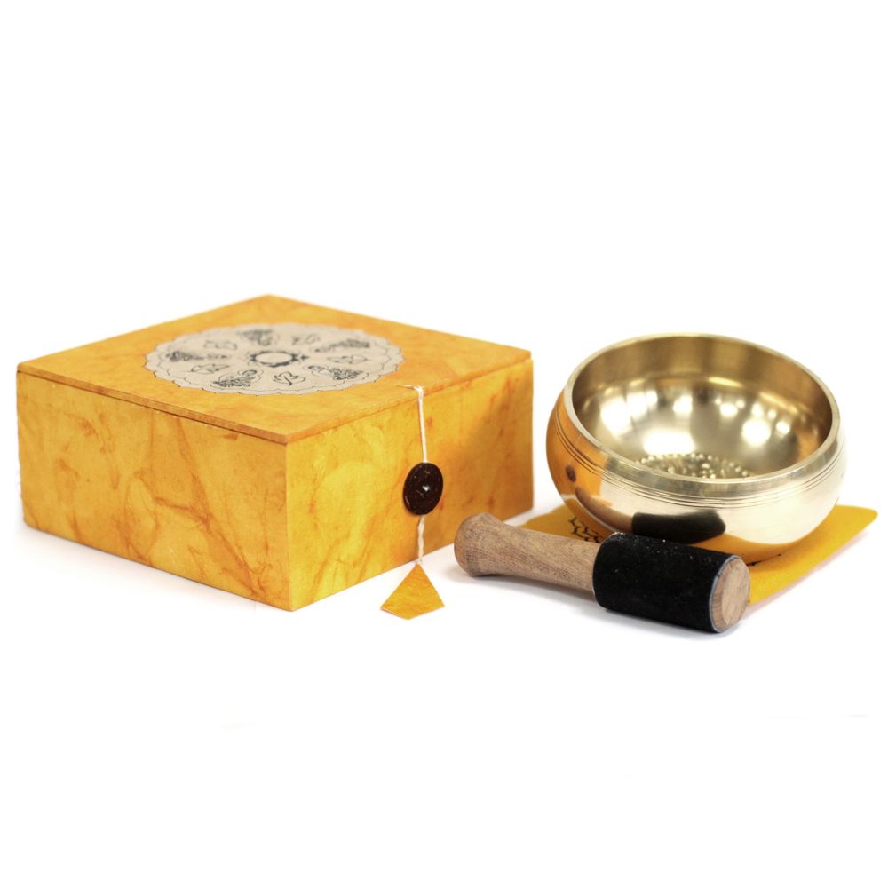 A brass singing bowl is placed on a cushion alongside a wooden mallet with a black handle, next to a decorative yellow box sealed with a string and wax seal.