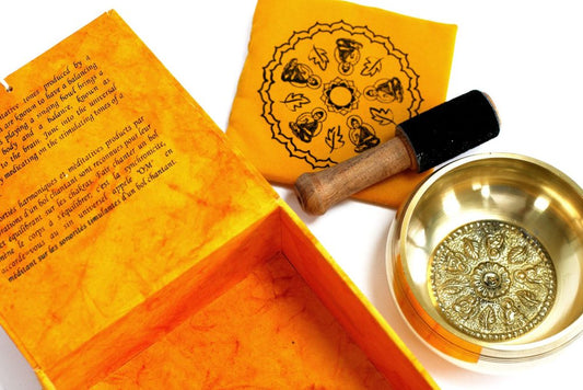 A brass singing bowl with intricate designs rests beside a wooden mallet and a patterned yellow cloth. Nearby, an open orange box displays text describing the bowl's harmonizing properties.