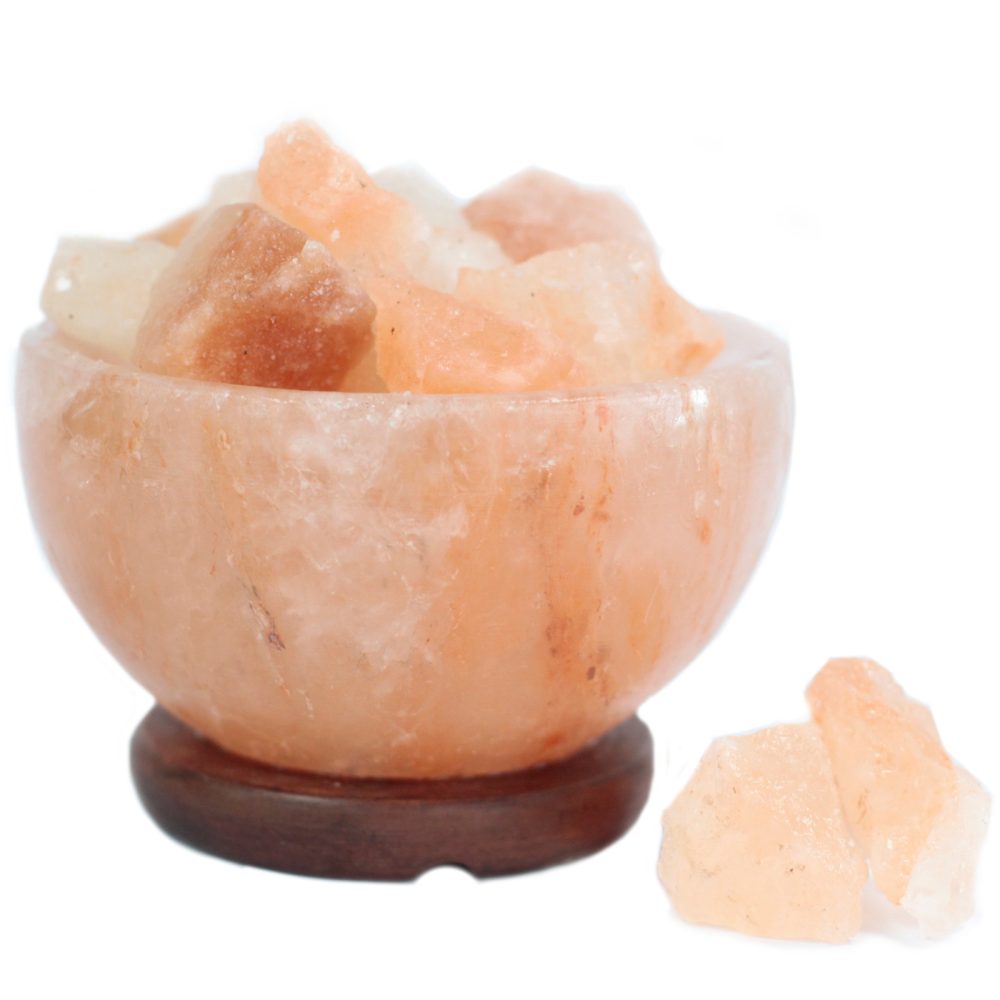 A round, pinkish Himalayan salt lamp is filled with large salt chunks, resting on a dark wooden base. Additional salt pieces lie beside it on a white background.
