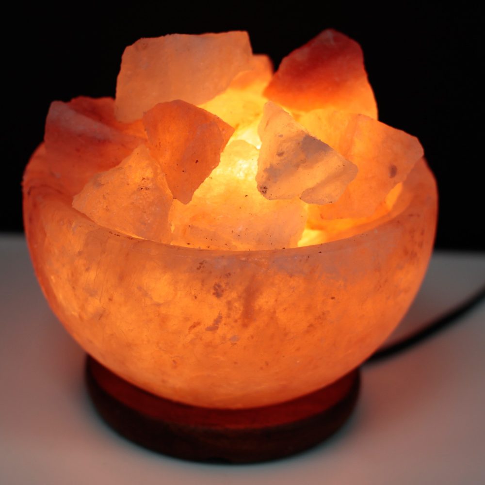 A glowing bowl of orange Himalayan salt crystals sits on a wooden base, illuminated from within, creating a warm, ambient light in a darkened setting.