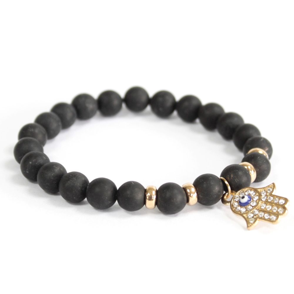 A beaded bracelet consisting of round, black stones interspersed with small gold cylinders and featuring a jeweled, gold Hamsa hand charm sits on a white background.