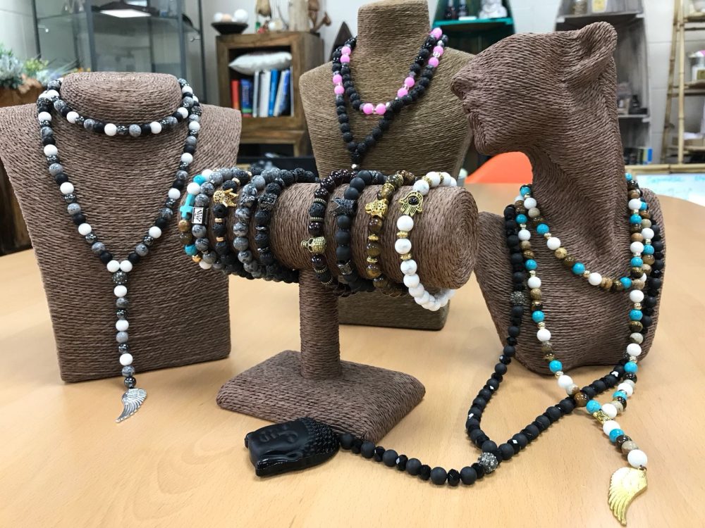 Jewelry is displayed on tan busts and a cylindrical stand on a light brown table. The jewelry includes beaded necklaces and bracelets in black, white, gray, gold, and turquoise colors.