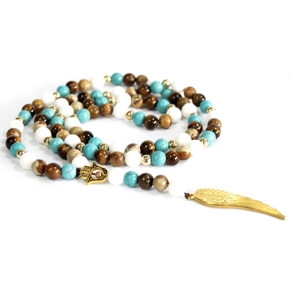 A mala necklace lies coiled and extended on a white surface. It is constructed with white, brown, and turquoise beads, gold spacers, a Hamsa charm, and a golden feather pendant.