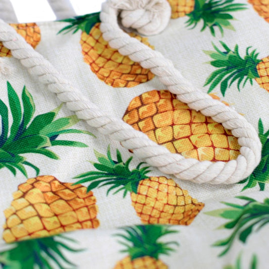 A tote bag lies, displaying printed yellow pineapples with green leaves on an off-white background, and a white twisted rope handle in the foreground.