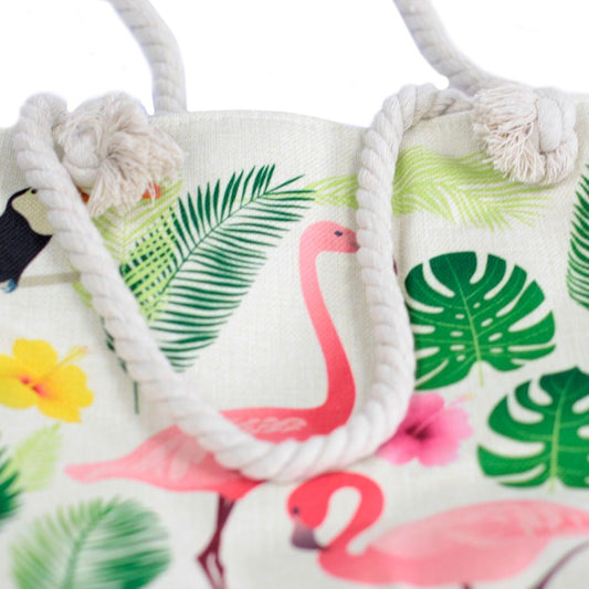 A tote bag with rope handles displays tropical illustrations, featuring pink flamingos, toucans, palm fronds, and hibiscus flowers, all on a white background.