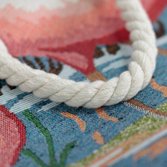 A thick white rope lays curved on a patterned, textured surface with pink, green, and blue tones, possibly depicting a flamingo and vegetation.