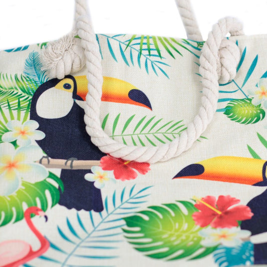 A canvas tote bag, with rope handles, shows printed images of toucans perched on branches, surrounded by tropical leaves, hibiscus flowers, and a flamingo.