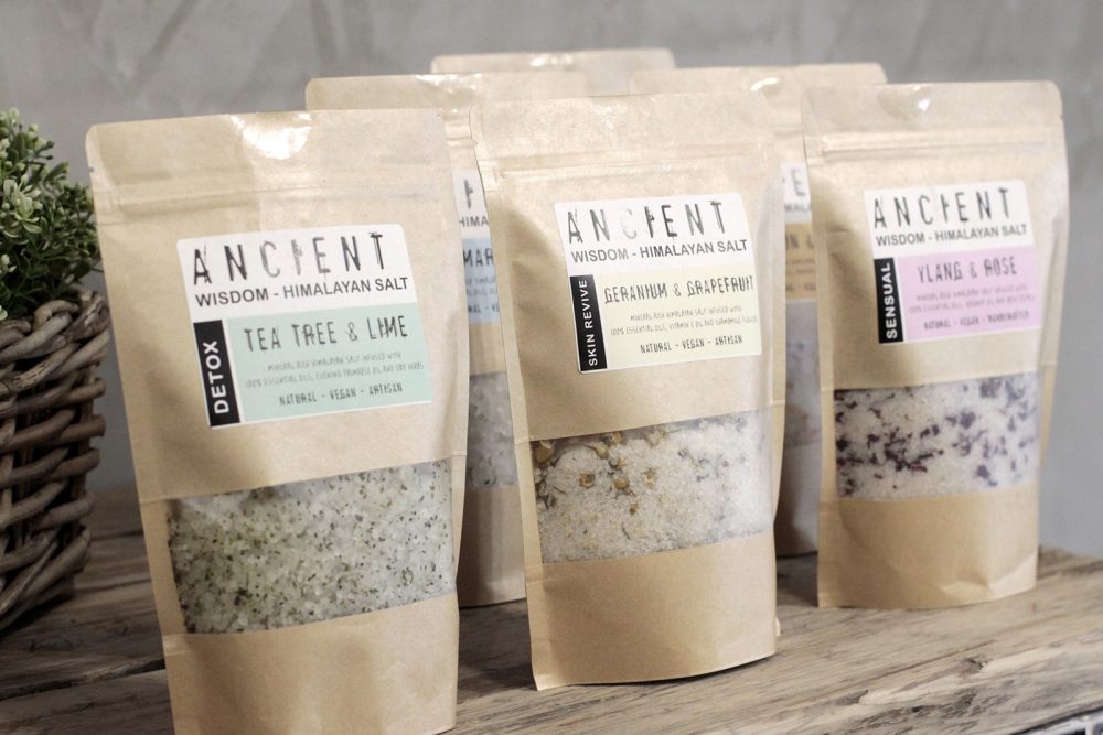 Several kraft paper bags, labeled "ANCIENT WISDOM - HIMALAYAN SALT," display different scents: "TEA TREE & LIME," "GERANIUM & GRAPEFRUIT," and "YLANG & ROSE," and sit on a wooden surface near a small plant.