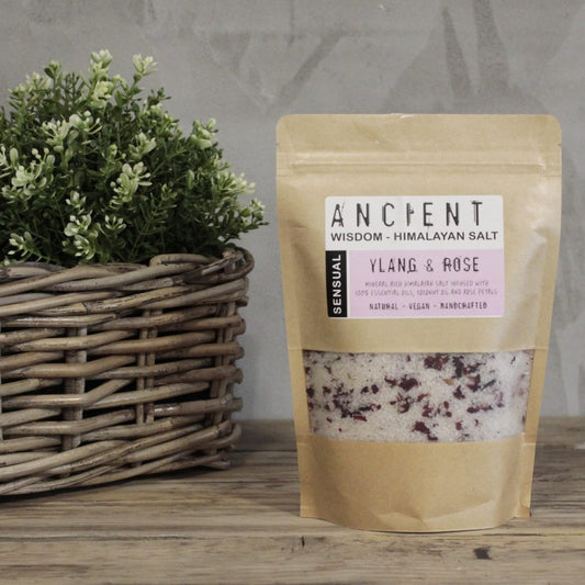 Bagged bath salts sit next to a potted plant on a wooden surface. The bag’s label reads: “ANCIENT WISDOM - HIMALAYAN SALT SENSUAL YLANG & ROSE MINERAL RICH HIMALAYAN SALT INFUSED WITH 100% ESSENTIAL OILS, COCONUT OIL AND ROSE PETALS NATURAL - VEGAN - HANDCRAFTED”.