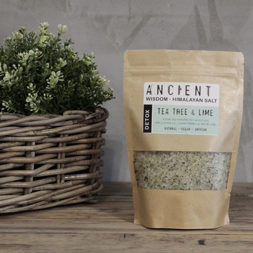 A bag of "ANCIENT Wisdom - Himalayan Salt," labeled "TEA TREE & LIME" and "DETOX," sits on a wooden surface. A wicker basket holding a green plant is adjacent to it.