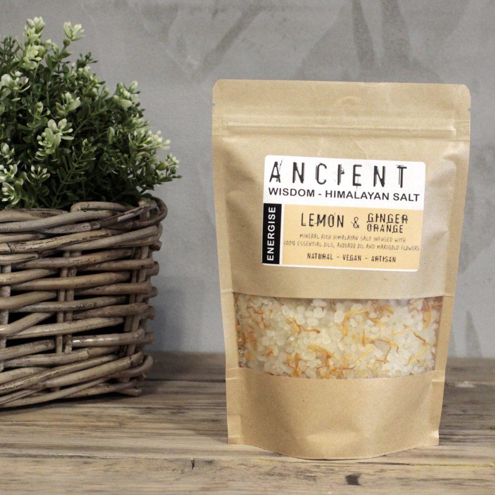 A packet of "ANCIENT WISDOM - HIMALAYAN SALT" stands on a wooden table, containing visible salt and orange bits. It is labeled "LEMON & GINGER ORANGE." A woven basket with a green plant sits beside it.