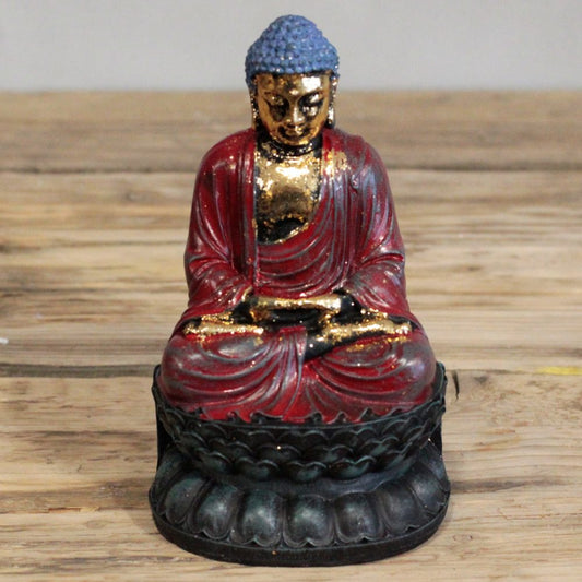 A small Buddha statue sits in meditation with eyes closed, draped in red robes with gold accents, on a lotus base. It's positioned on a wooden surface, creating a serene setting.