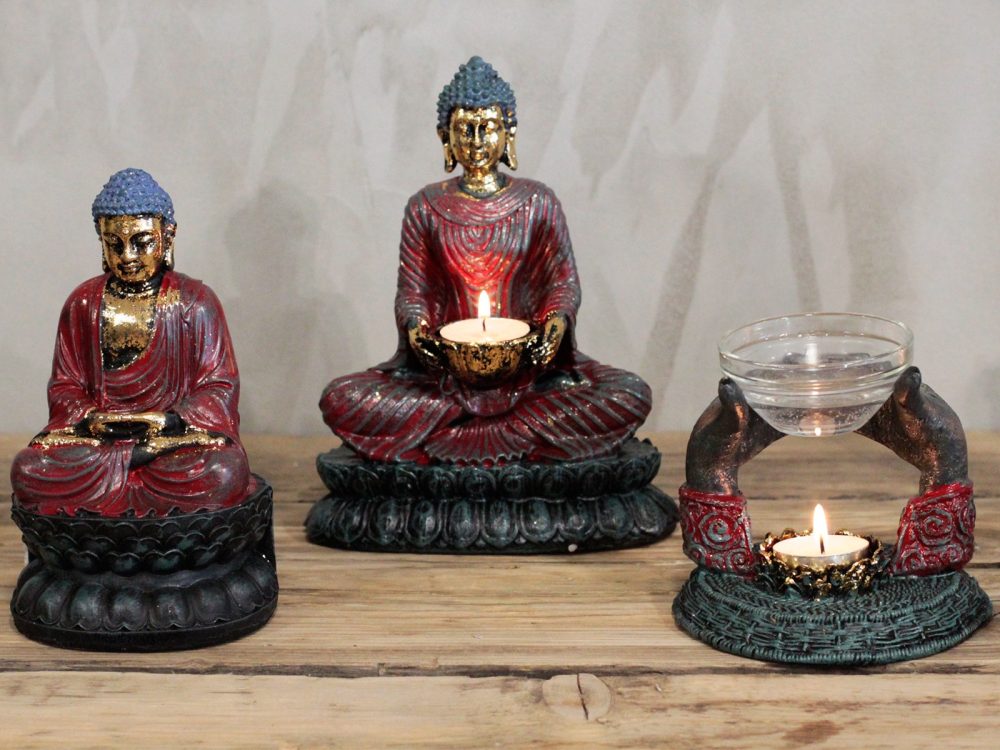 Two seated Buddha statues, one holding a lit tea candle, flank a two-armed decorative stand with a glass bowl and lit tealight on a wooden table against a gray wall.