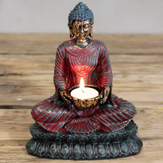 Buddha statue holds a lit tealight in a small bowl, seated cross-legged on a lotus base, painted red and gold with blue green patina, placed on a rustic wooden tabletop.