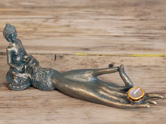 A metallic sculpture features a reclining Buddha figure beside an extended hand holding a small candle. The setting is a wooden surface with visible grains and textures.