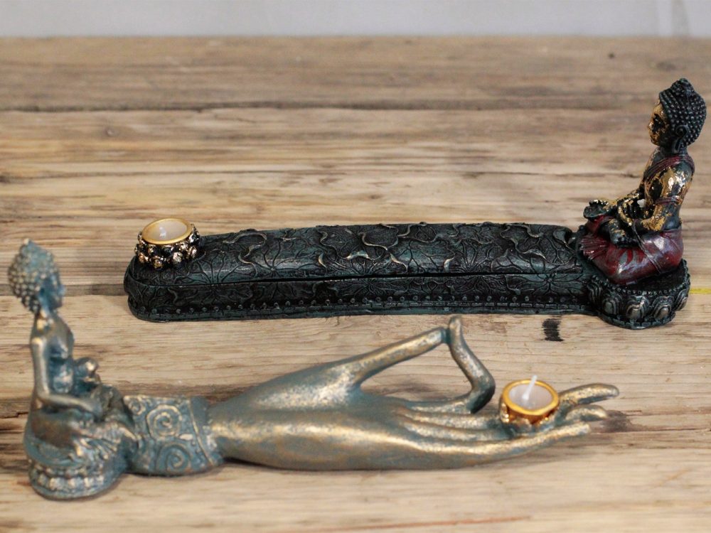 Two ornate Buddha-shaped candle holders rest on a wooden surface; one lies horizontally, depicting a reclining figure, while the other stands upright with a seated figure.