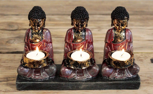Three Buddha statues, each holding a lit tealight candle, sit in a row on a wooden surface. Their red and gold tones create a warm, tranquil ambiance.