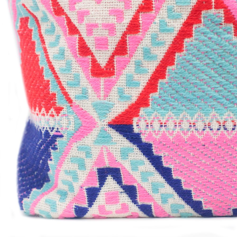 A woven bag displays colorful patterns. Geometric shapes interlock, rendered in blue, pink, red, and turquoise on an off-white background, against a plain white backdrop.