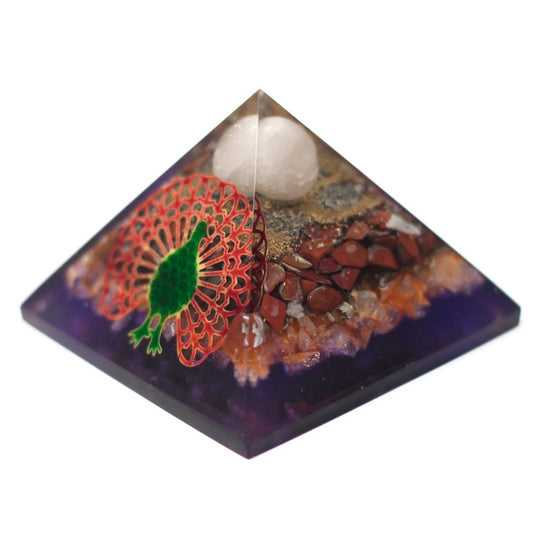 An orgone pyramid sits on a white surface; inside the pyramid are a white stone at the top, layered with red stones, gold flakes, and a green and red peacock design.
