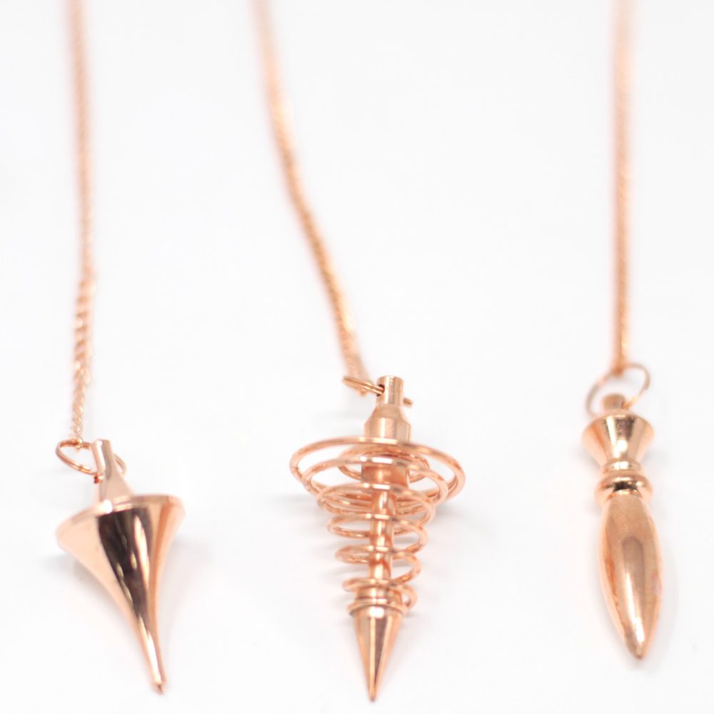 Three rose-gold pendulums with long chains hang in the center against a bright white background, each a different geometric shape.