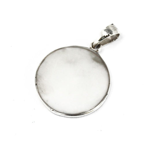A silver pendant, round with a shiny white center, lays on a white surface. The pendant's loop faces the upper right corner.
