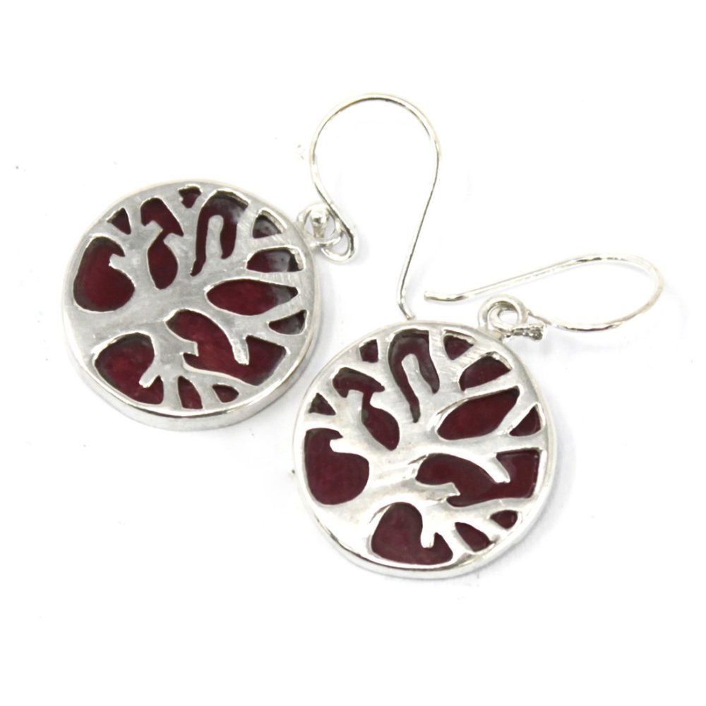Two silver tree-of-life earrings dangle. The tree design is cut out of the silver circles, revealing a burgundy-colored backing. They are displayed on a white surface.