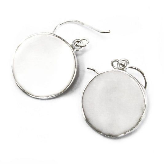 Two silver earrings with circular, opaque white stones rest on a bright white surface. Each earring has a curved hook for piercing.