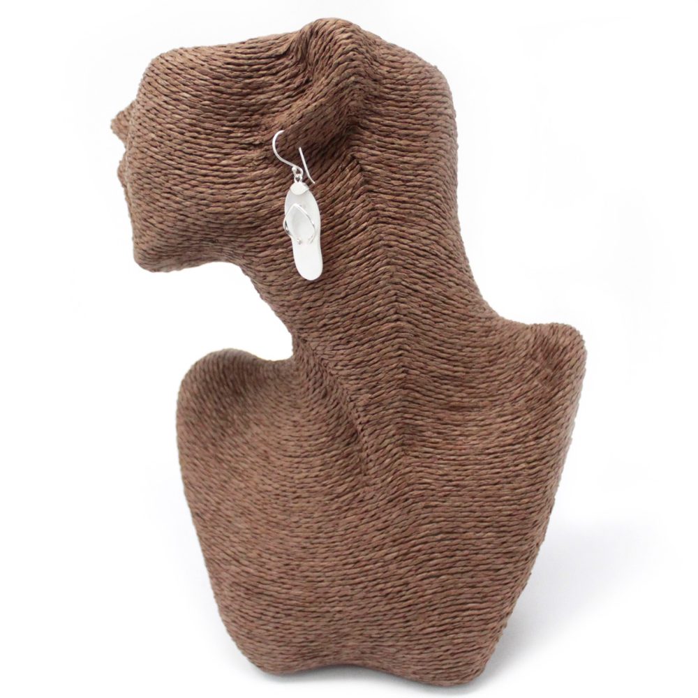 A silver earring shaped like a flip-flop dangles from the ear of a brown bust, that sits against a solid white background.