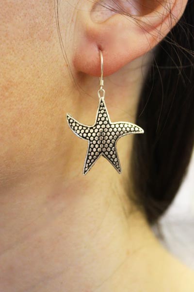 A silver starfish earring dangles from a pierced earlobe. The earring is textured with small dots, against a blurry background of skin and hair.