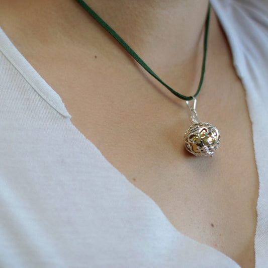 A silver filigree ball pendant hangs from a dark green cord around a person's neck. The person is wearing a white t-shirt.