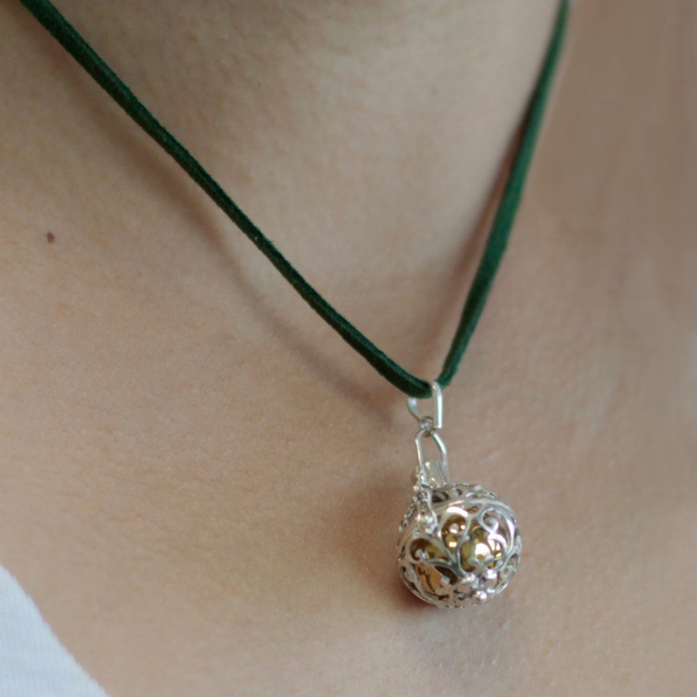 A silver, filigree sphere pendant hangs from a green cord around a person's neck. The pendant dangles against skin.