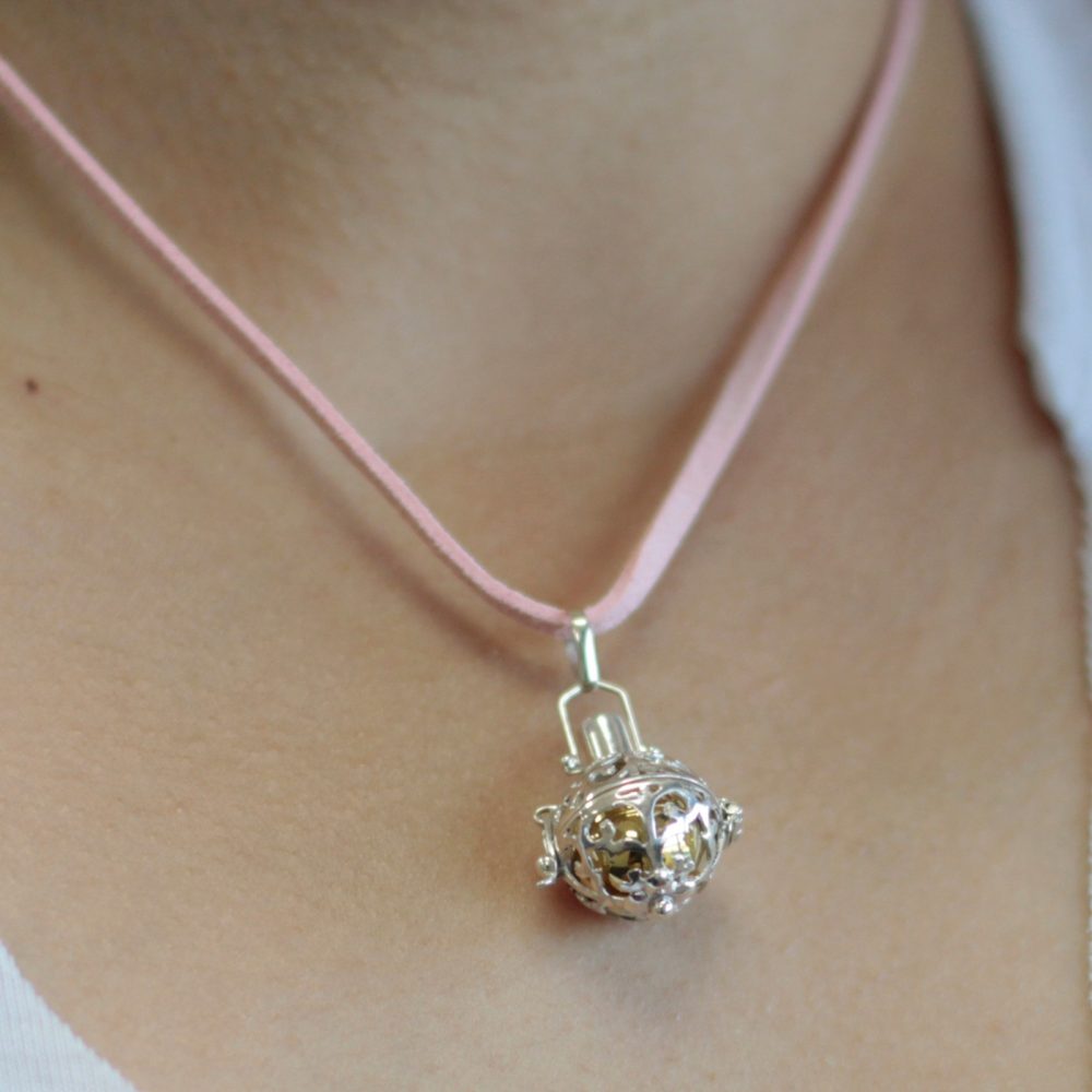 A silver pendant dangles from a light pink cord necklace against a light-skinned person’s neck. The pendant is a spherical cage containing a golden-colored ball inside.