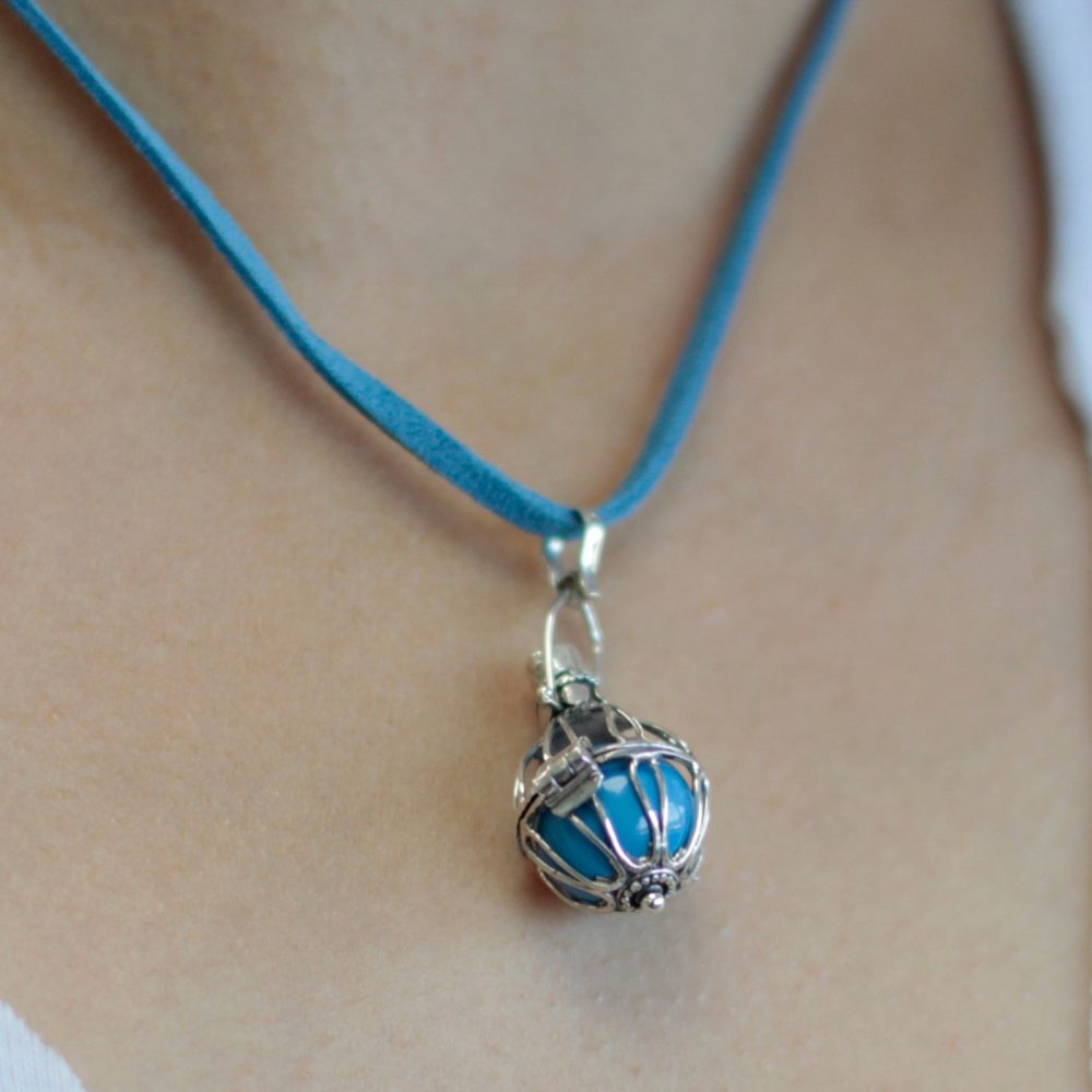 A silver cage pendant holds a turquoise stone, suspended from a blue suede cord. The necklace rests on a person’s fair skin and white clothing.