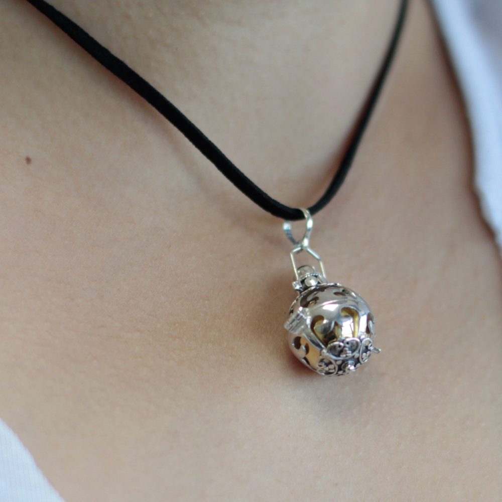 A silver pendant hangs from a black cord around a person's neck. The spherical pendant has decorative cutouts.